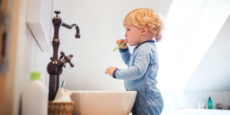 curly-haired toddler in a striped onesie stands in front of mirror and sink to brush teeth